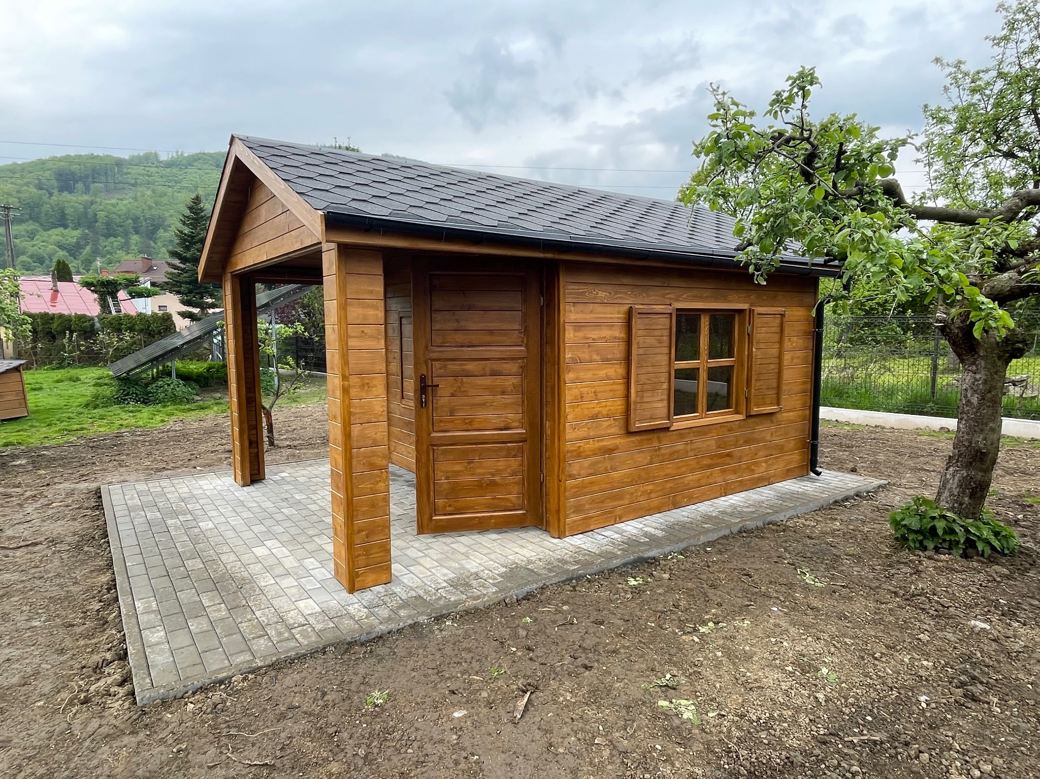 Drewniana altana z otwartym gankiem i oknem, pomalowana na brązowo, stoi na wyłożonym kostką placu. W tle zielone wzgórza i drzewa.