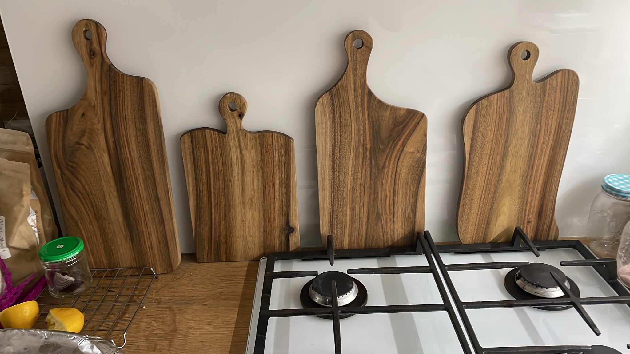 Cztery drewniane deski do krojenia o różnych kształtach, ustawione na blacie obok kuchenki gazowej, w tle biała ściana. Widoczne akcesoria kuchenne.