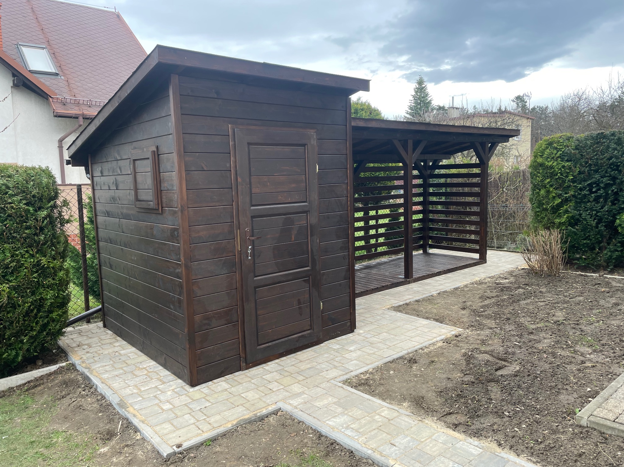 Ciemnobrązowa, drewniana altana z zamykanym pomieszczeniem gospodarczym, połączona z otwartą pergolą, na tle pochmurnego nieba i zieleni.