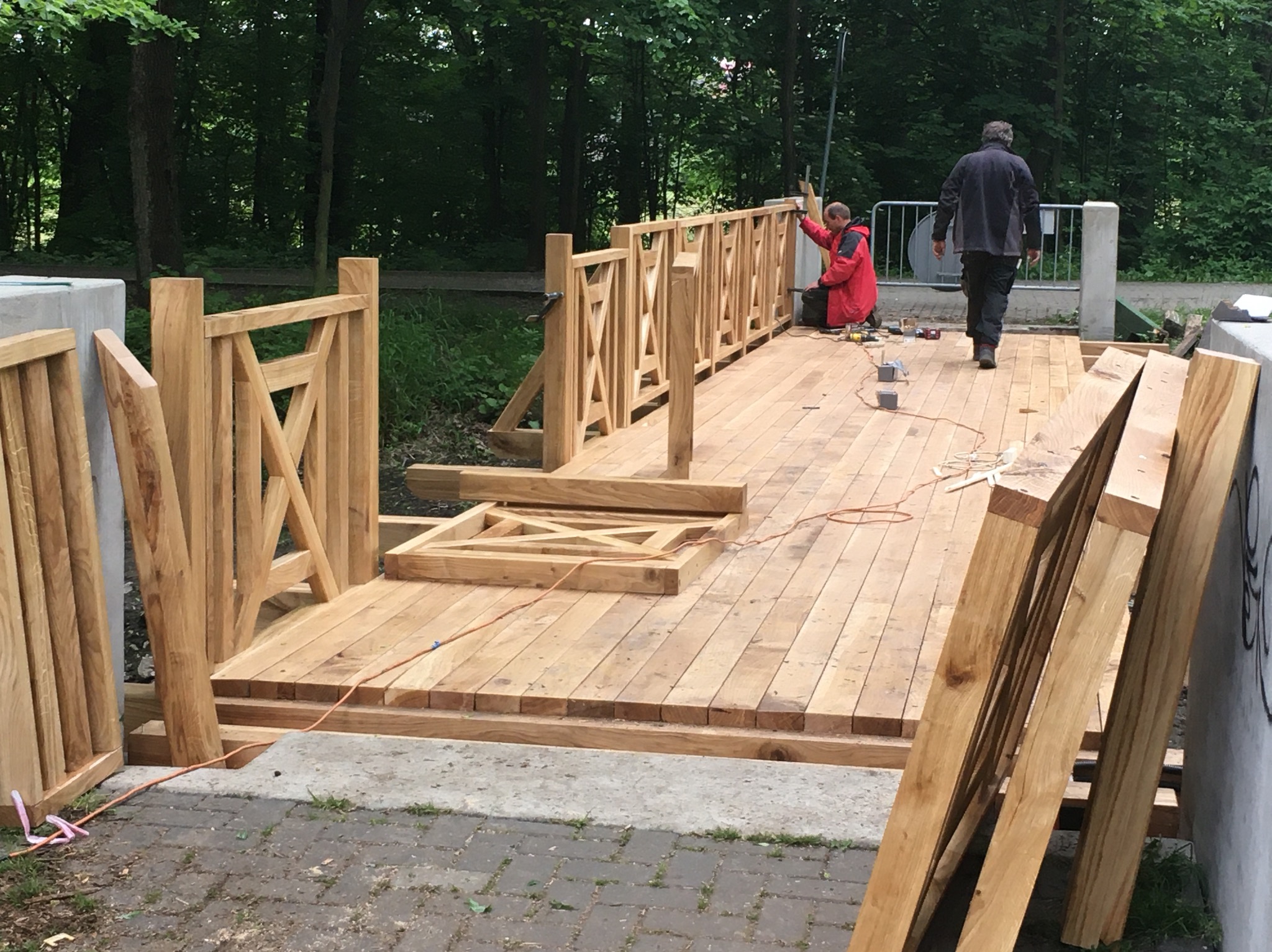 Montaż drewnianego mostku z balustradami w parku, dwóch mężczyzn przy pracy, jeden w czerwonej kurtce, otoczenie zieleni. Elementy balustrad oparte o betonowe słupki.
