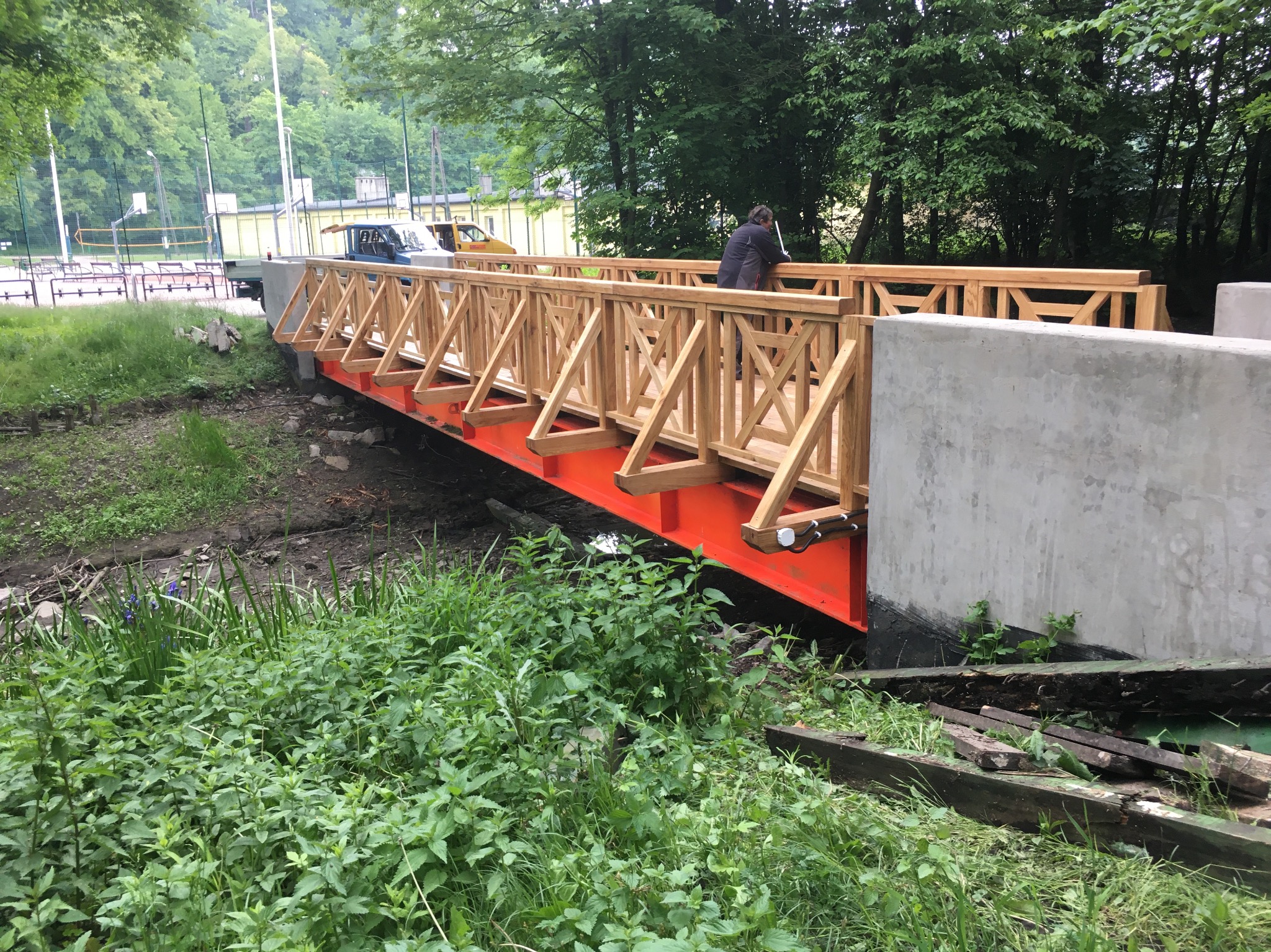 Nowy, drewniany mostek z pomarańczową konstrukcją stalową, z osobą opartą o balustradę. W tle zieleń, boisko i budynek. Kąt ujęcia z dołu.