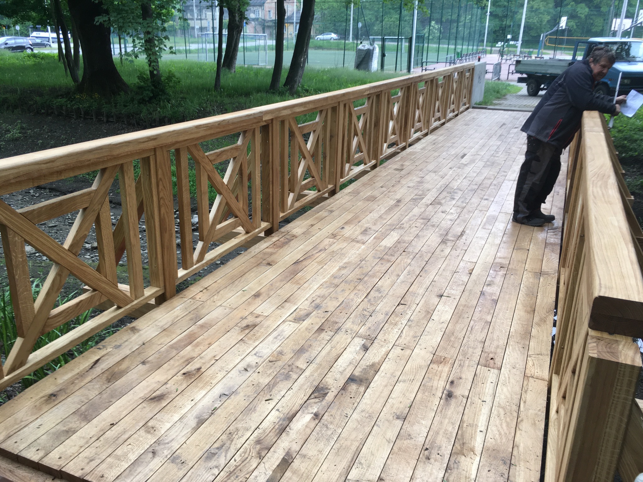 Nowa, drewniana kładka z balustradami o wzorze krzyżowym, na tle zieleni i boiska. Mężczyzna pochyla się nad dokumentami, opierając się o barierkę.