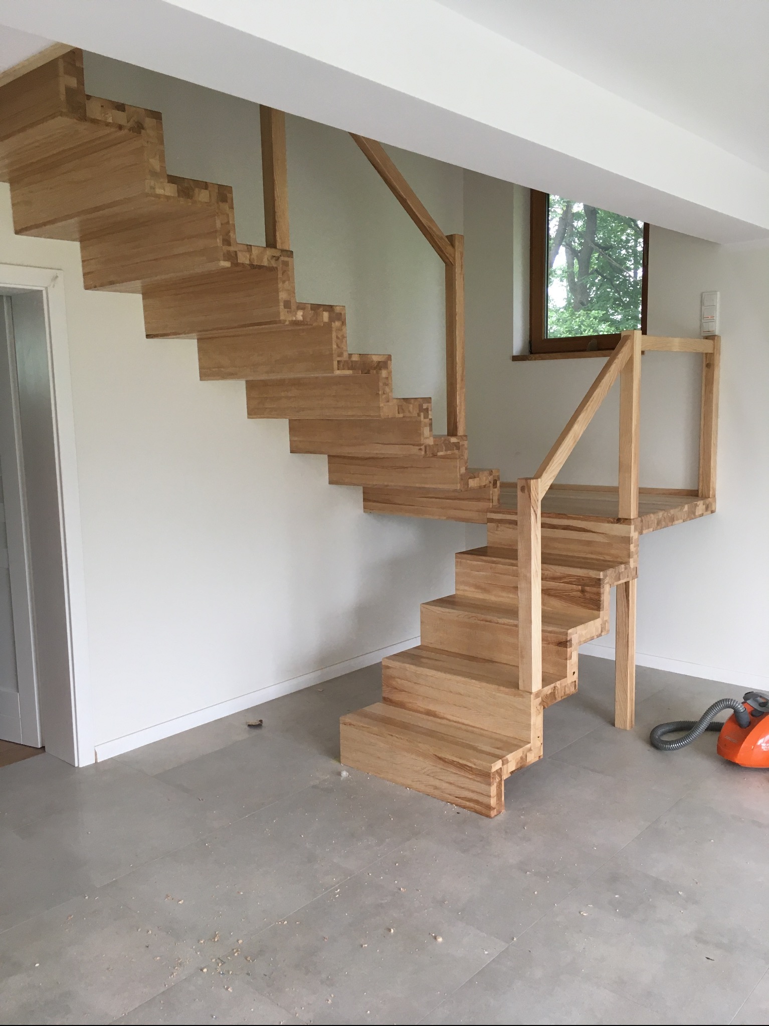 Surowe, drewniane schody z balustradą w jasnym wnętrzu. Widoczne niedoskonałości i pył po obróbce, pomarańczowy odkurzacz. Schody bez wykończenia, w trakcie montażu.