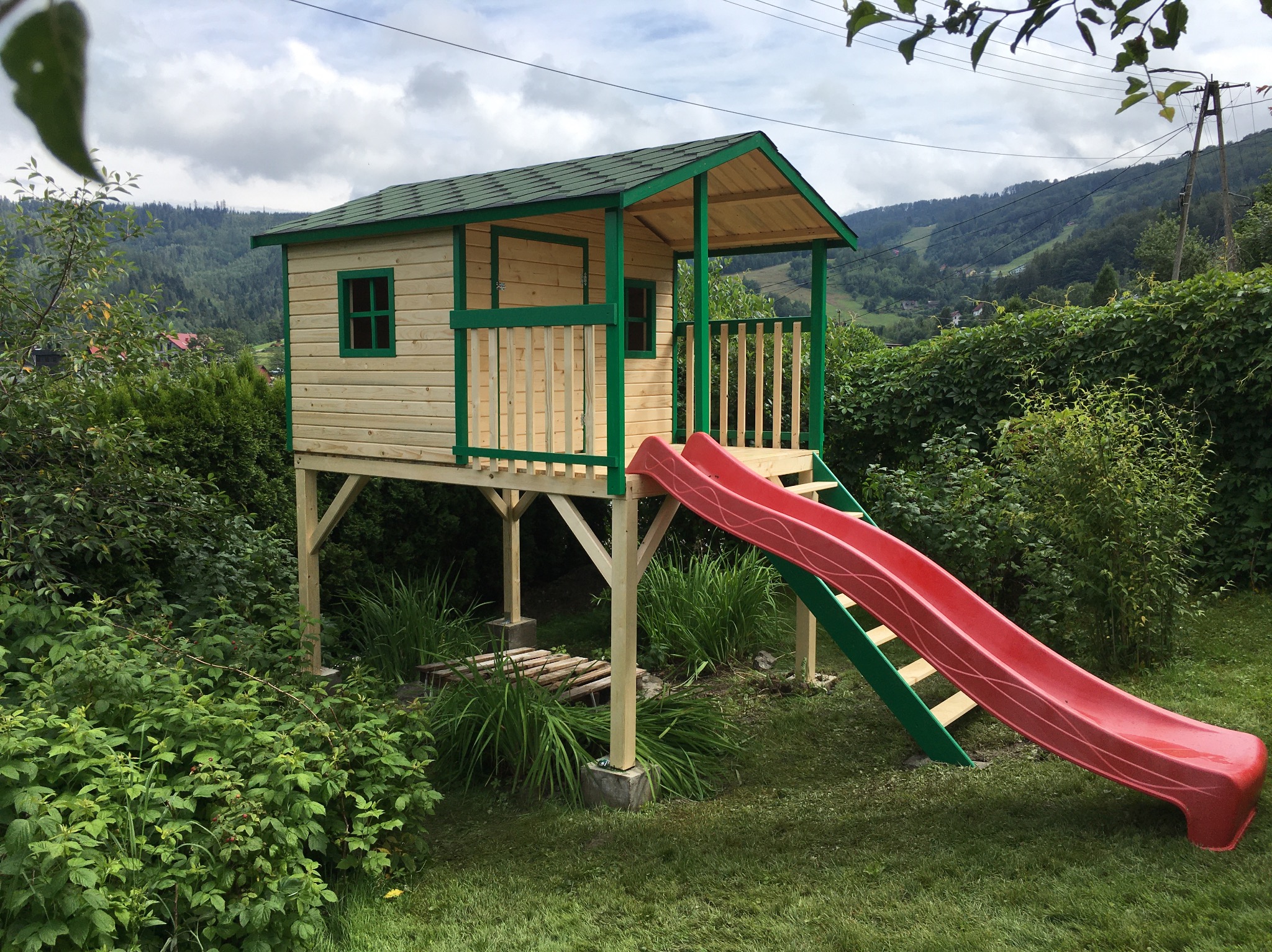 Drewniany domek dla dzieci na podwyższeniu z czerwoną zjeżdżalnią, otoczony zielenią. Widok na góry w tle, dom z zielonymi akcentami.