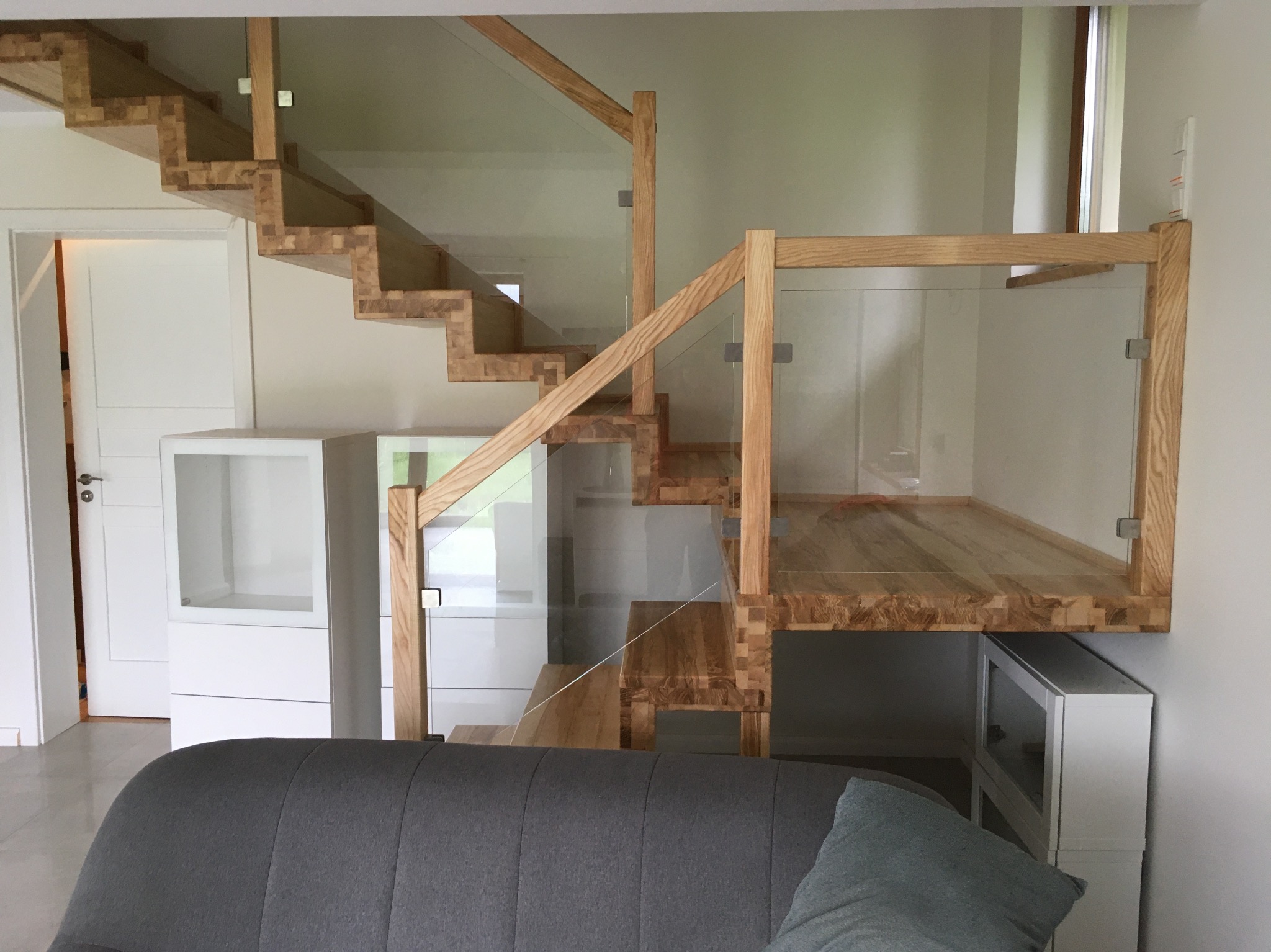 Nowoczesne schody drewniane z szklaną balustradą, widok z dołu. Stopnie z litego drewna, minimalistyczny design, jasne wnętrze. Widoczna część sofy.