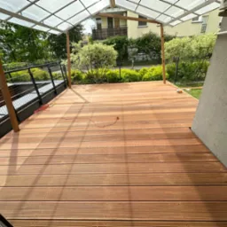 Zadaszony taras z desek kompozytowych w odcieniach brązu, otoczony zielenią i elementami architektury. Widoczny fragment ściany budynku i balustrada.