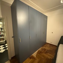 Struktura drewna - Duża, szara szafa wnękowa z złotymi uchwytami, dopasowana do podłogi z parkietu w jodełkę. W tle widoczny fragment pokoju z czarno-białą podłogą.