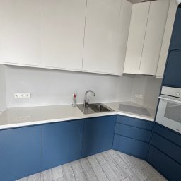 Struktura drewna - Nowoczesna kuchnia z białymi szafkami górnymi i granatowymi dolnymi. Blat jasny, zlew stalowy. Minimalistyczny design, widoczne gniazdka elektryczne i fragment podłogi.