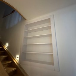 Struktura drewna - Wnęka z półkami na ścianie klatki schodowej, oświetlenie punktowe schodów. Białe półki w ramie, widok od dołu, drewniane schody.