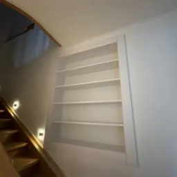 Wnęka z półkami na ścianie klatki schodowej, oświetlenie punktowe schodów. Białe półki w ramie, widok od dołu, drewniane schody.