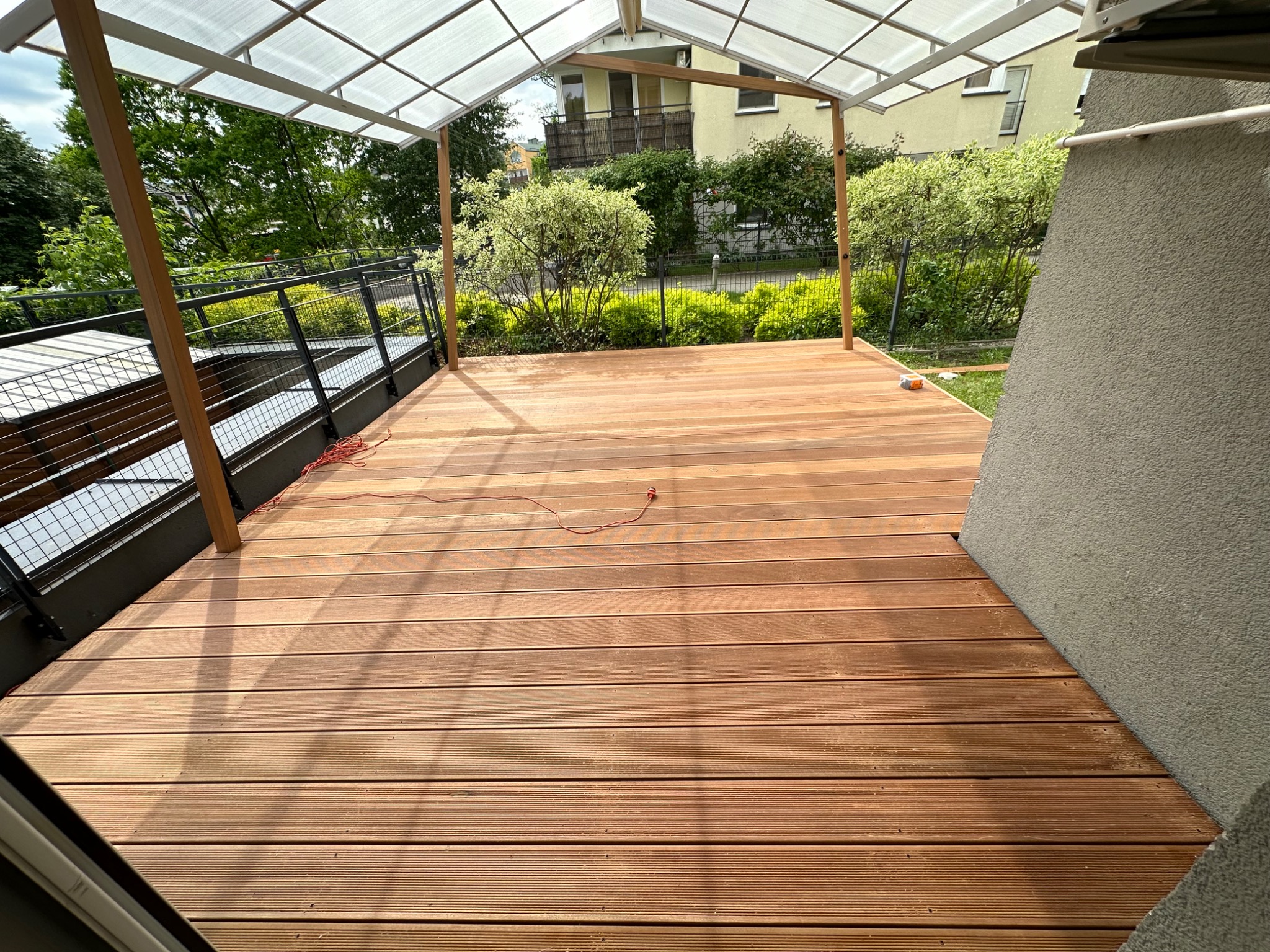 Zadaszony taras z desek kompozytowych w odcieniach brązu, otoczony zielenią i elementami architektury. Widoczny fragment ściany budynku i balustrada.