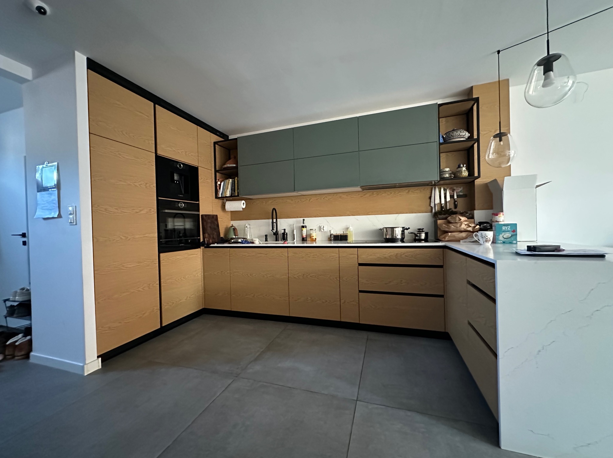 Nowoczesna kuchnia z drewnianymi frontami, szarymi szafkami wiszącymi i wyspą z białym blatem. Oświetlenie punktowe i szare płytki podłogowe.