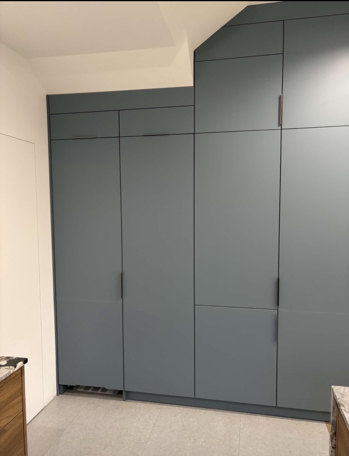 Nowoczesna, szara zabudowa szaf wnękowych z minimalistycznymi uchwytami, dopasowana do skosu dachu. Widoczny fragment blatu kuchennego.