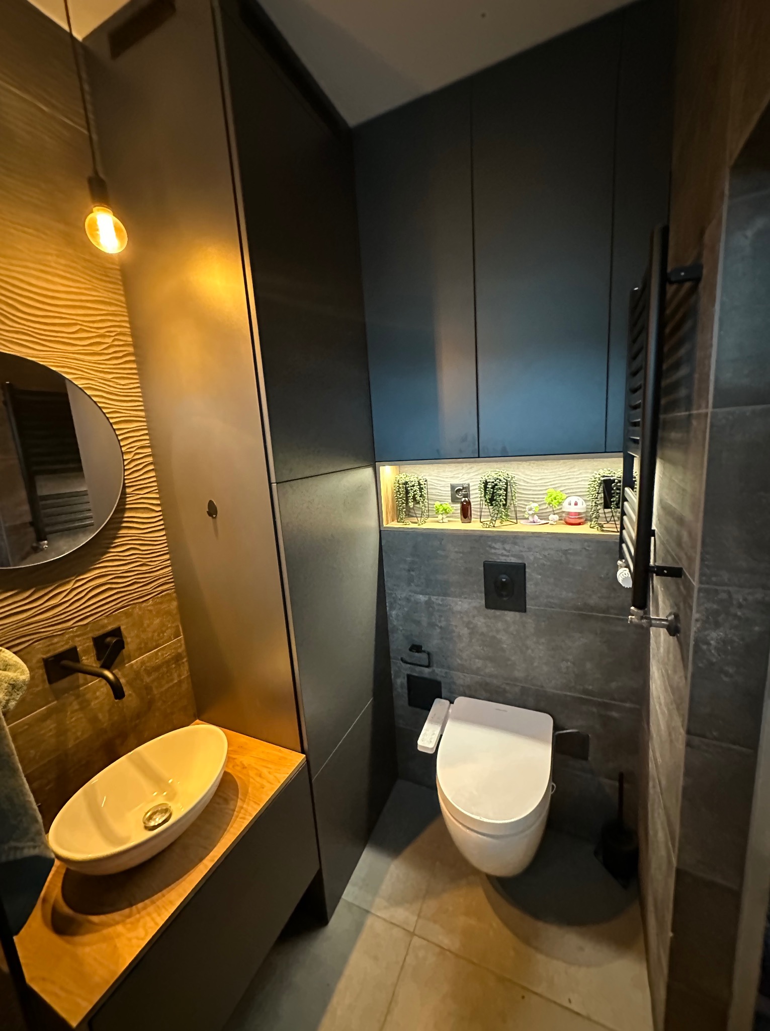 Nowoczesna łazienka w ciemnych kolorach z podświetlaną półką nad toaletą i umywalką nablatową. W tle widoczne lustro i dekoracyjna ściana.