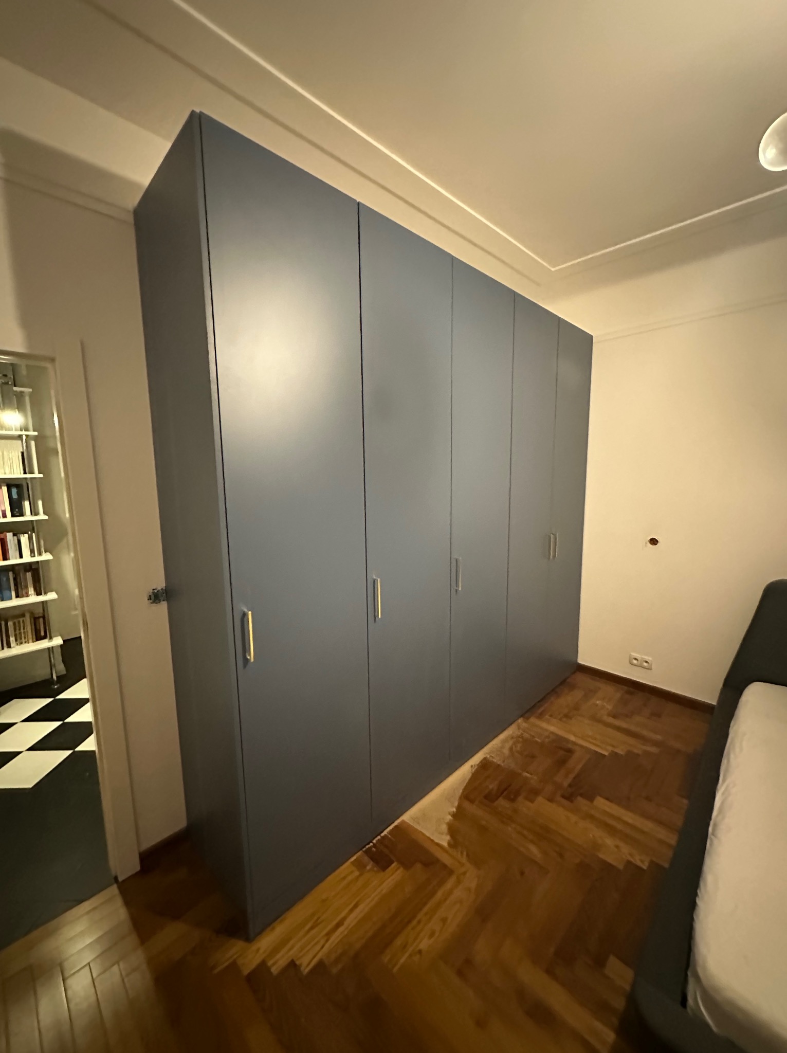 Duża, szara szafa wnękowa z złotymi uchwytami, dopasowana do podłogi z parkietu w jodełkę. W tle widoczny fragment pokoju z czarno-białą podłogą.