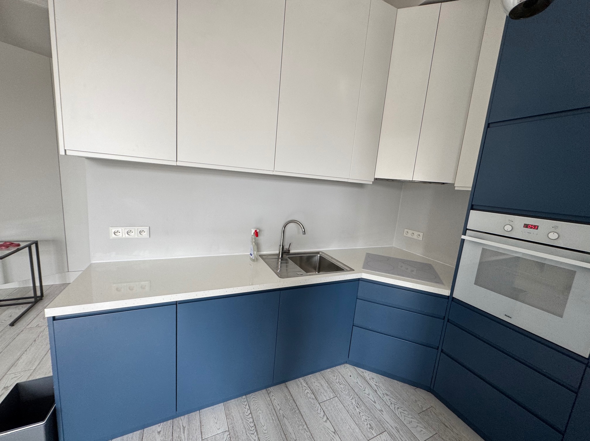 Nowoczesna kuchnia z białymi szafkami górnymi i granatowymi dolnymi. Blat jasny, zlew stalowy. Minimalistyczny design, widoczne gniazdka elektryczne i fragment podłogi.