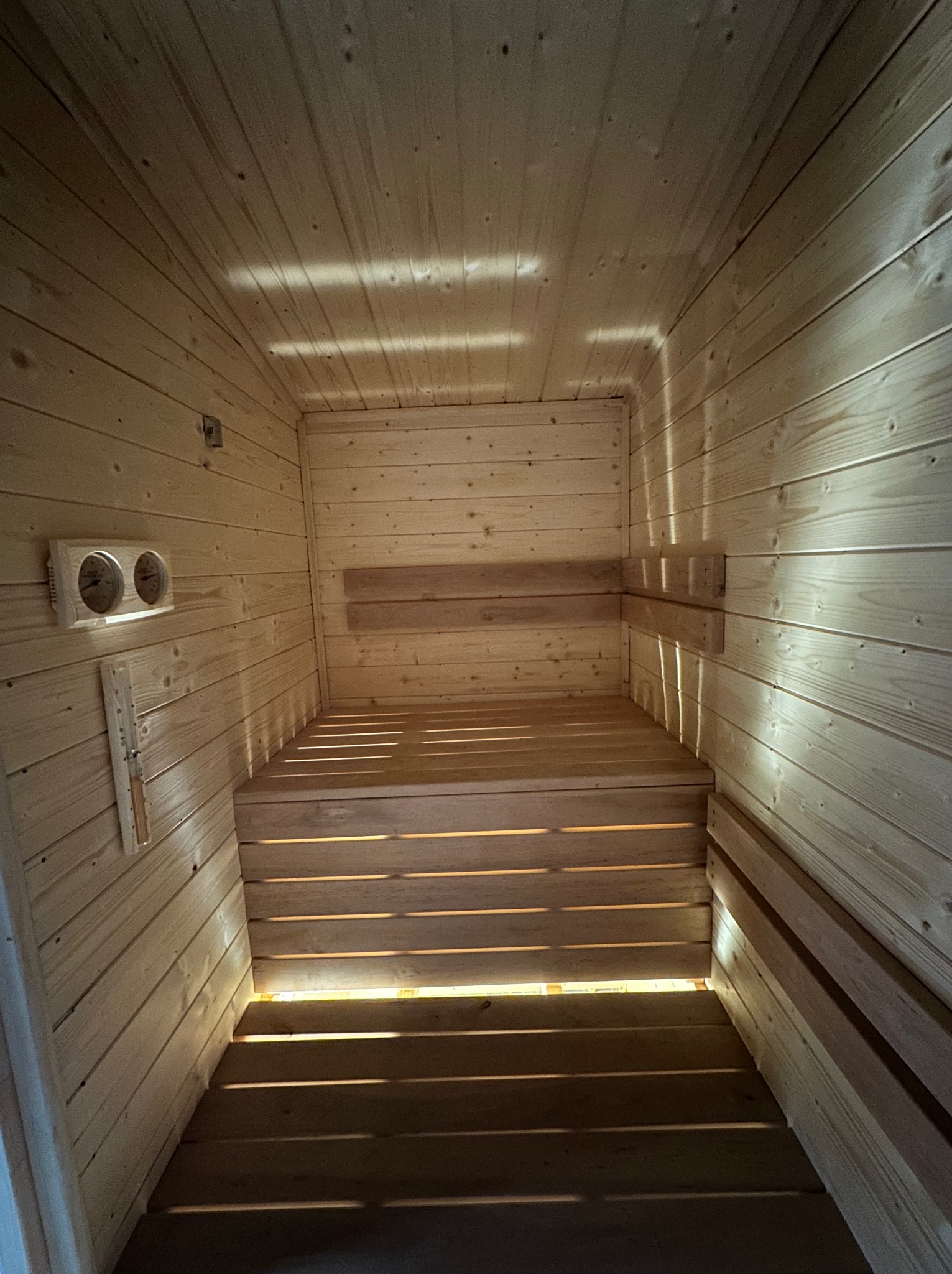 Wnętrze sauny z jasnego drewna, widok z dołu na ławy i ścianę z termometrem. Naturalne światło podkreśla strukturę drewna.