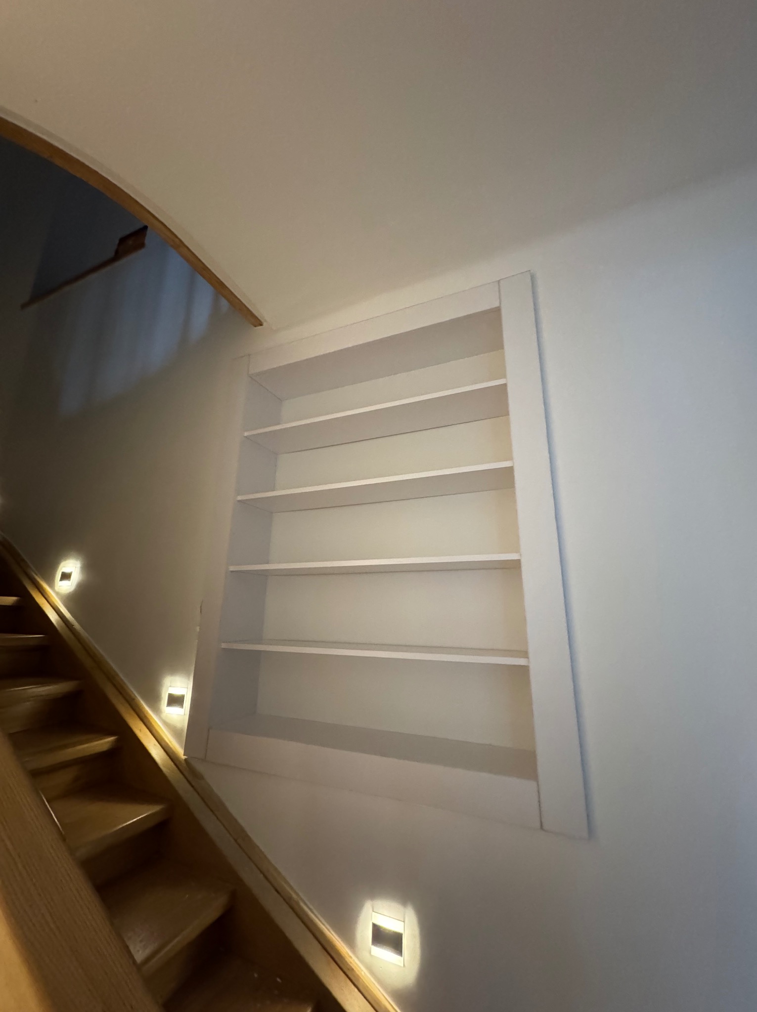 Wnęka z półkami na ścianie klatki schodowej, oświetlenie punktowe schodów. Białe półki w ramie, widok od dołu, drewniane schody.