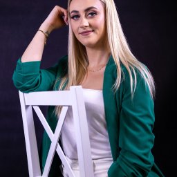 Bartosz Wejs Studio - usługi fotograficzne i filmowe oraz montaż sieci teletechnicznych - Portret kobiety z blond włosami, w zielonej marynarce, siedzącej na białym krześle na ciemnym tle. Uśmiechnięta, z ręką opartą o głowę, patrzy w obiektyw.