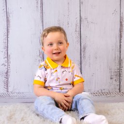 Bartosz Wejs Studio - usługi fotograficzne i filmowe oraz montaż sieci teletechnicznych - Uśmiechnięty chłopiec w koszulce z motywem budowlanym siedzi na puszystym dywanie na tle drewnianej ściany. Jasne, naturalne światło.