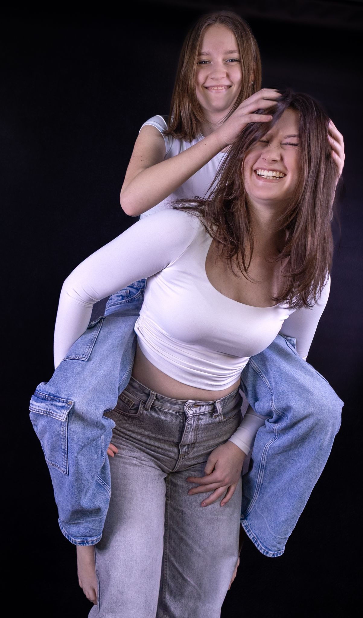 Dwie roześmiane nastolatki w białych bluzkach i jeansach na czarnym tle: jedna na barana, druga czesze ją rękami, wyraźne emocje i spontaniczność.