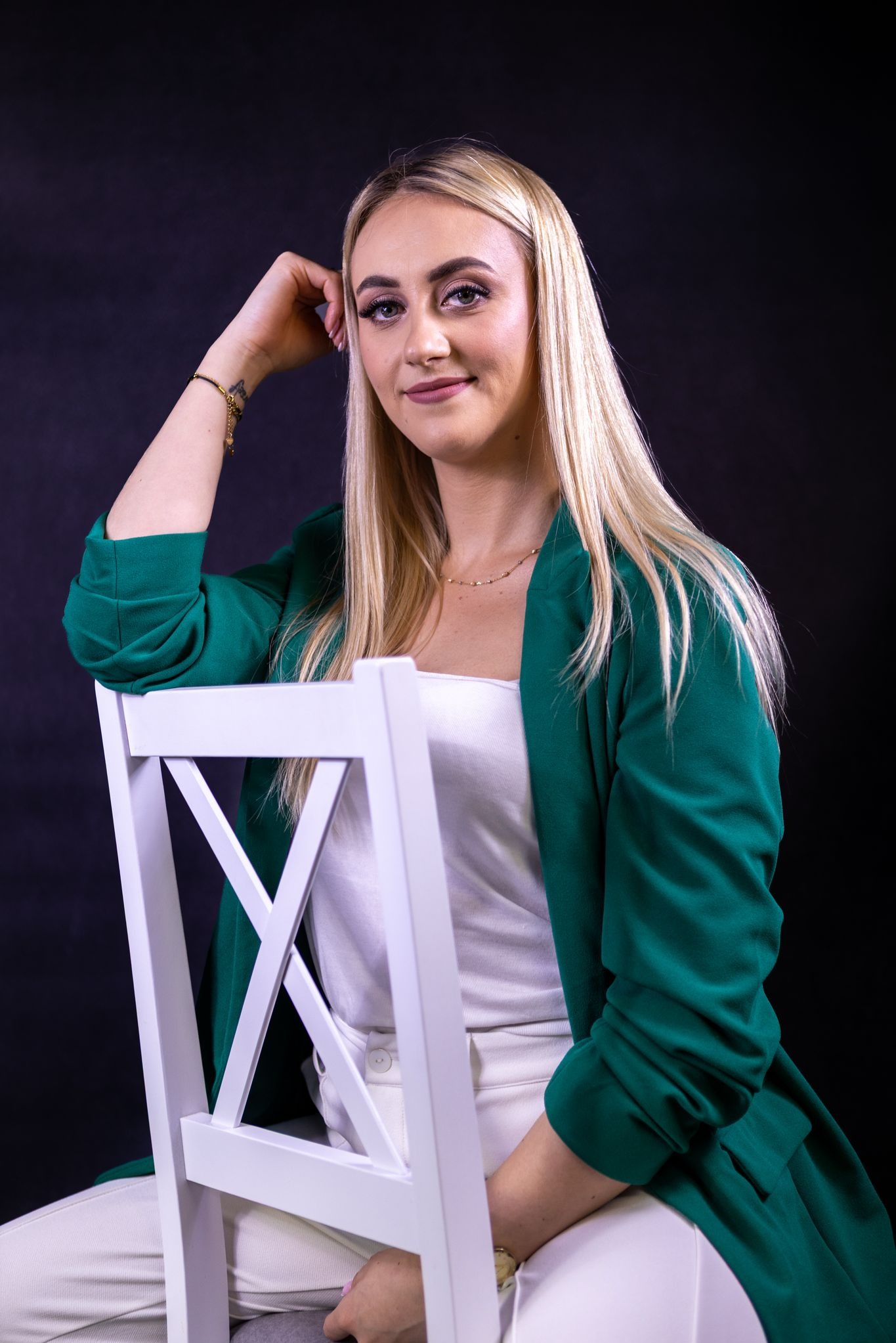 Portret kobiety z blond włosami, w zielonej marynarce, siedzącej na białym krześle na ciemnym tle. Uśmiechnięta, z ręką opartą o głowę, patrzy w obiektyw.