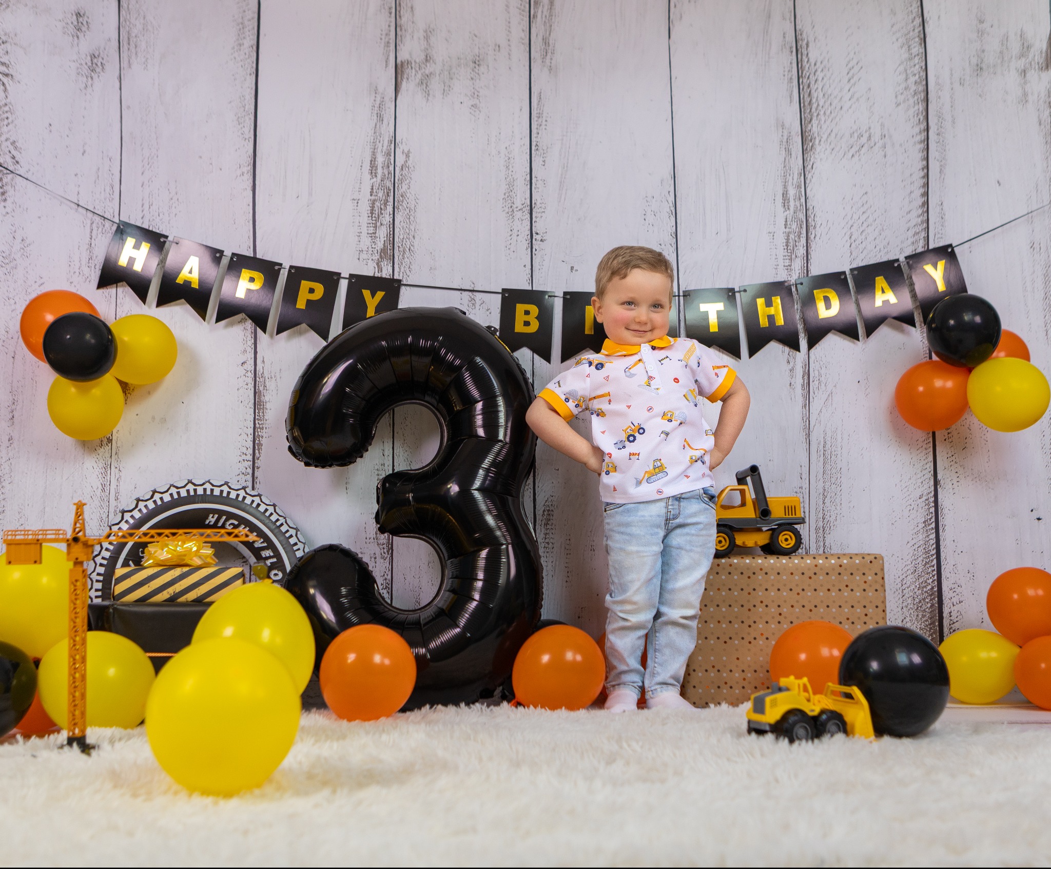 Uśmiechnięty chłopiec w koszulce z motywem budowlanym na tle urodzinowej dekoracji z balonami i zabawkami, w tematyce budowlanej. Balon w kształcie cyfry 3.