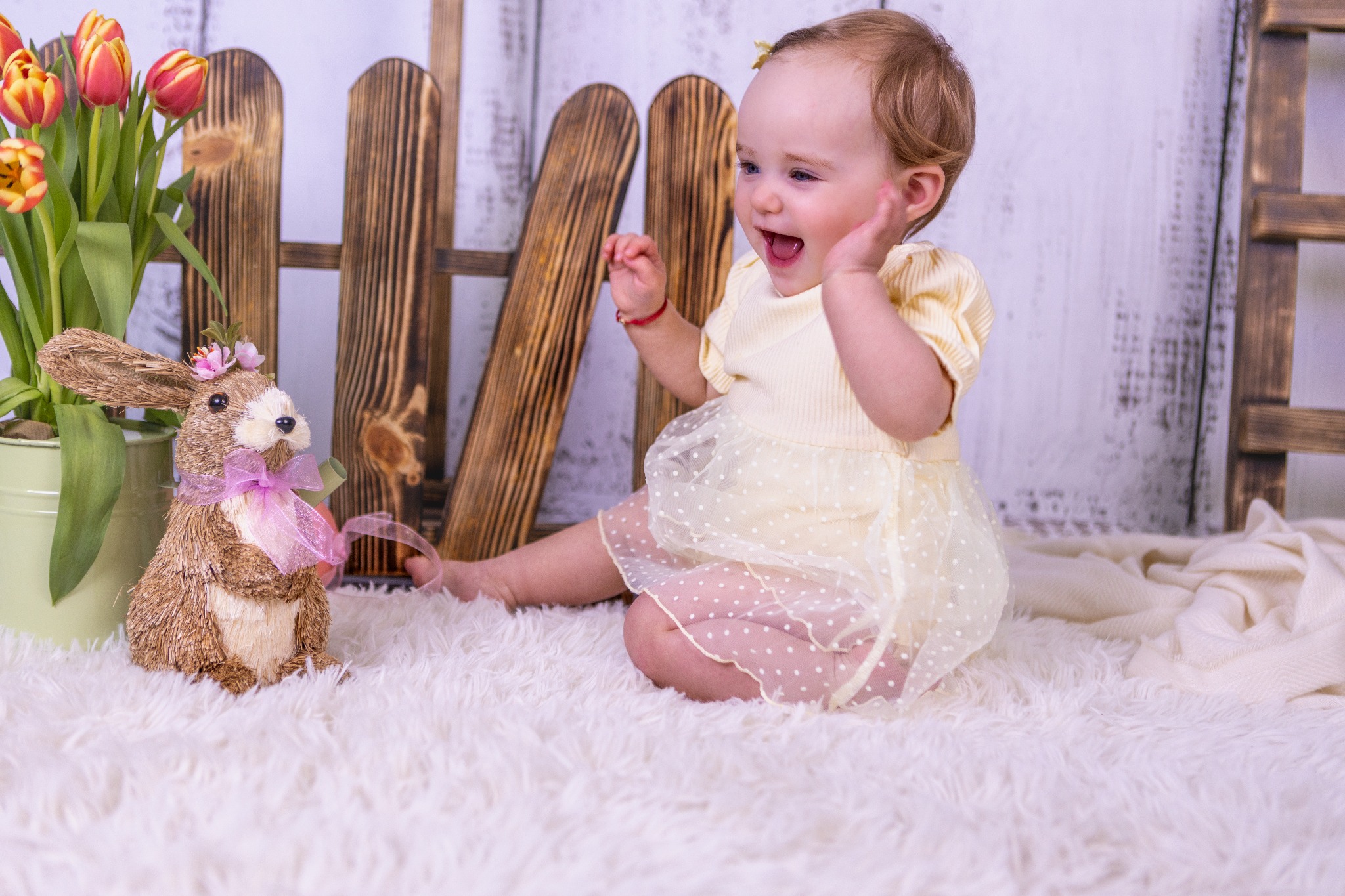 Radosna dziewczynka w żółtej sukience bawi się obok dekoracyjnego zajączka i tulipanów na tle drewnianego płotku. Jasne, pastelowe kolory.