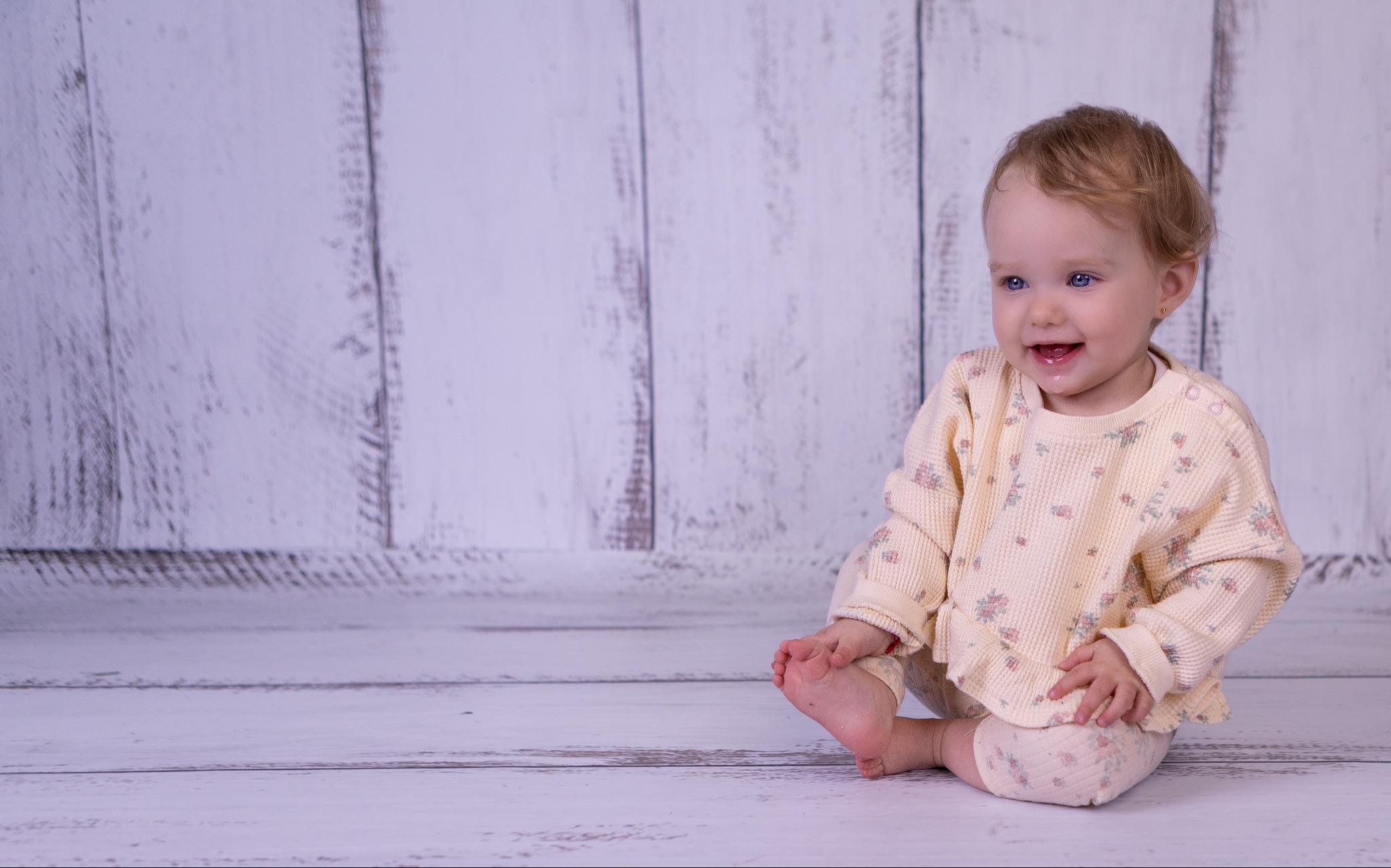 Uśmiechnięta, roczna dziewczynka z blond włosami i niebieskimi oczami siedzi na drewnianej podłodze w jasnym ubranku w kwiatki na tle białej ściany z desek.