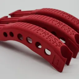 Czerwone, łukowate uchwyty z napisem 'PRESS', ułożone jeden za drugim. Wyraźna tekstura i okrągłe otwory w każdym elemencie. Detal wykonany prawdopodobnie z tworzywa.