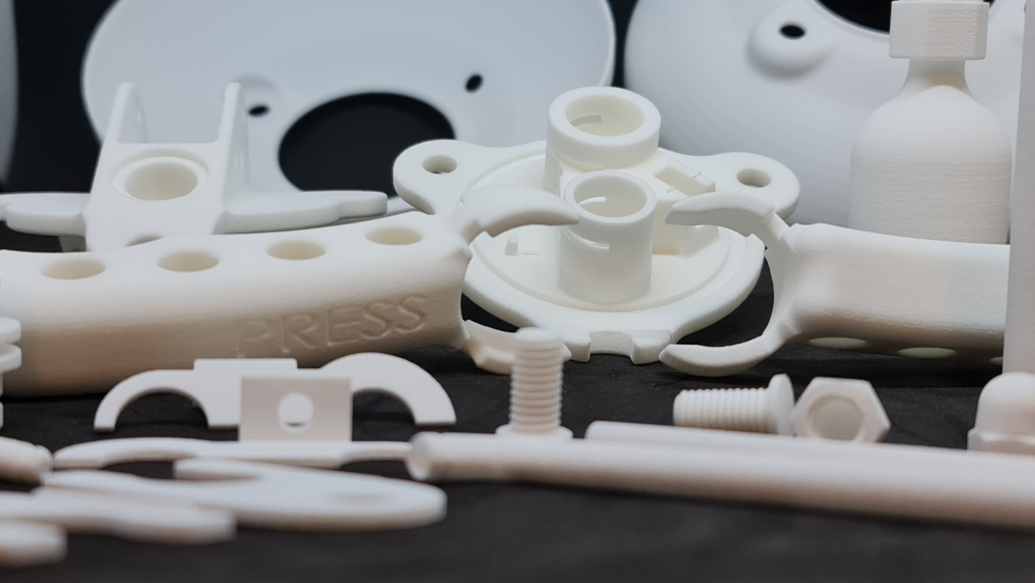 Białe, precyzyjnie wykonane elementy z tworzywa sztucznego, w tym śruby i nakrętki, ułożone na czarnym tle. Widoczne detale i faktura materiału.