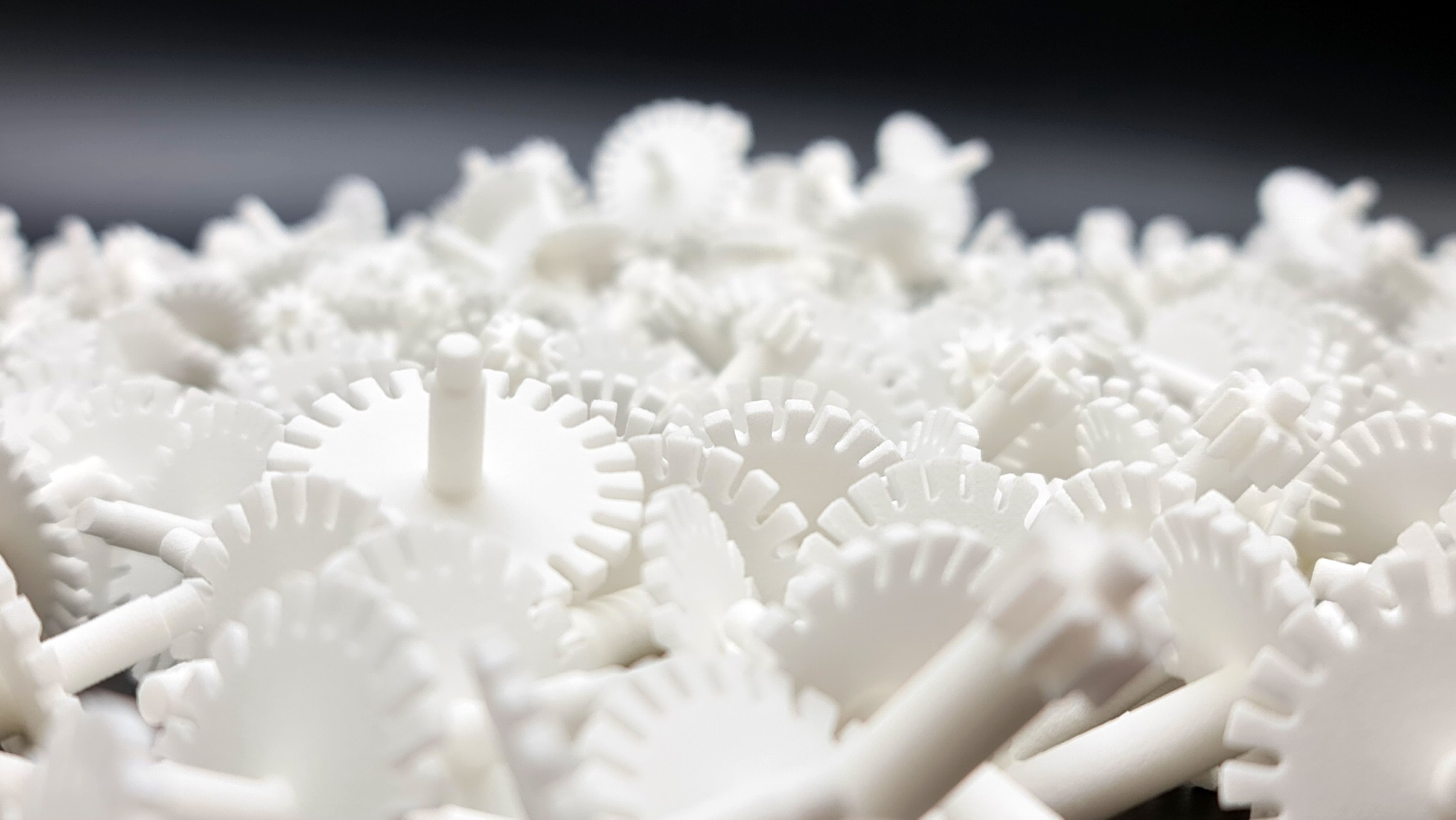 Białe, plastikowe koła zębate z trzpieniami w dużym skupisku, oświetlone z góry, na ciemnym tle. Detal precyzyjnego wykonania.