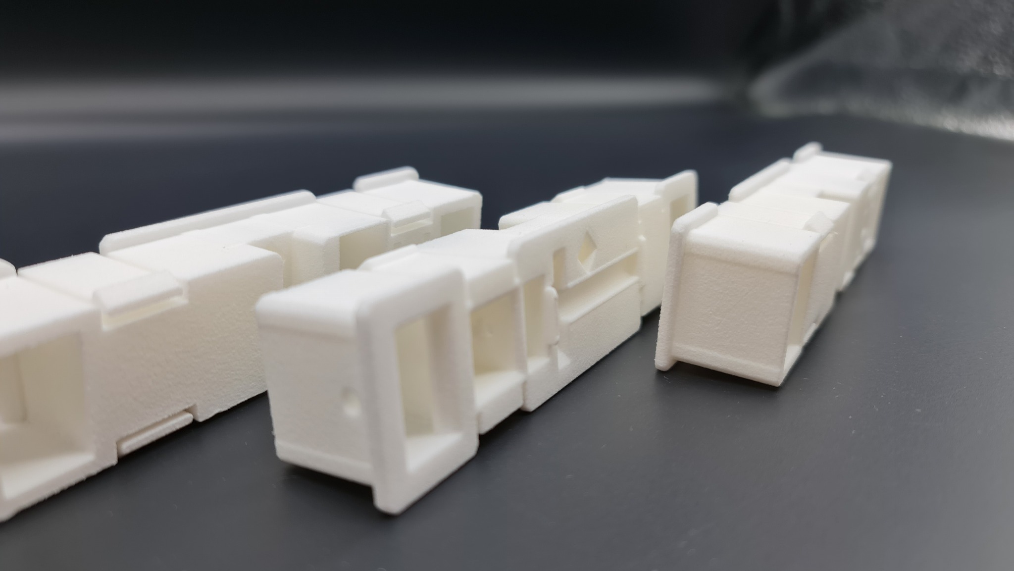 Białe, modułowe elementy z tworzywa sztucznego, prawdopodobnie wydrukowane w 3D, ułożone na ciemnoszarym tle. Widoczna tekstura druku.