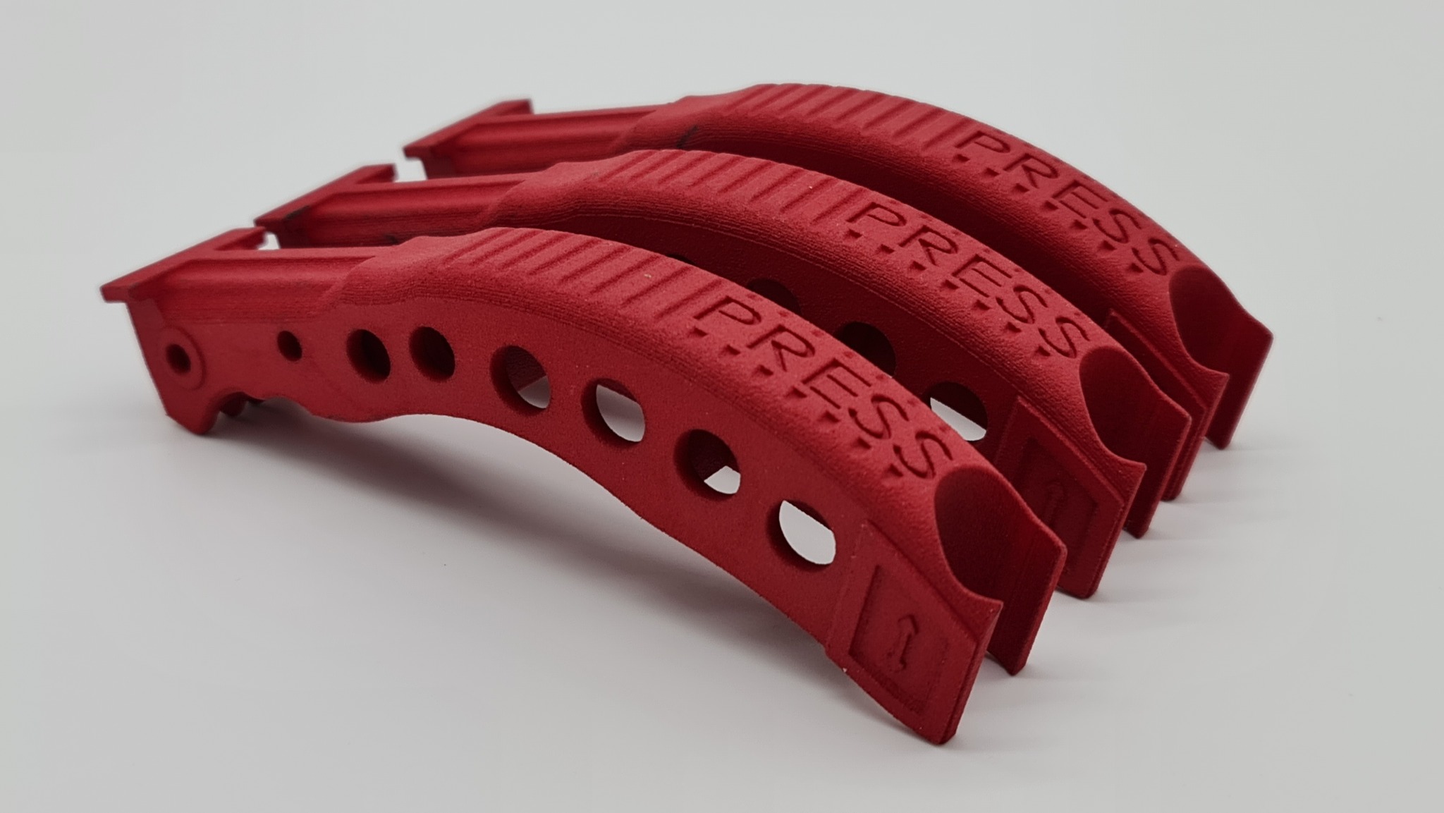 Czerwone, łukowate uchwyty z napisem 'PRESS', ułożone jeden za drugim. Wyraźna tekstura i okrągłe otwory w każdym elemencie. Detal wykonany prawdopodobnie z tworzywa.