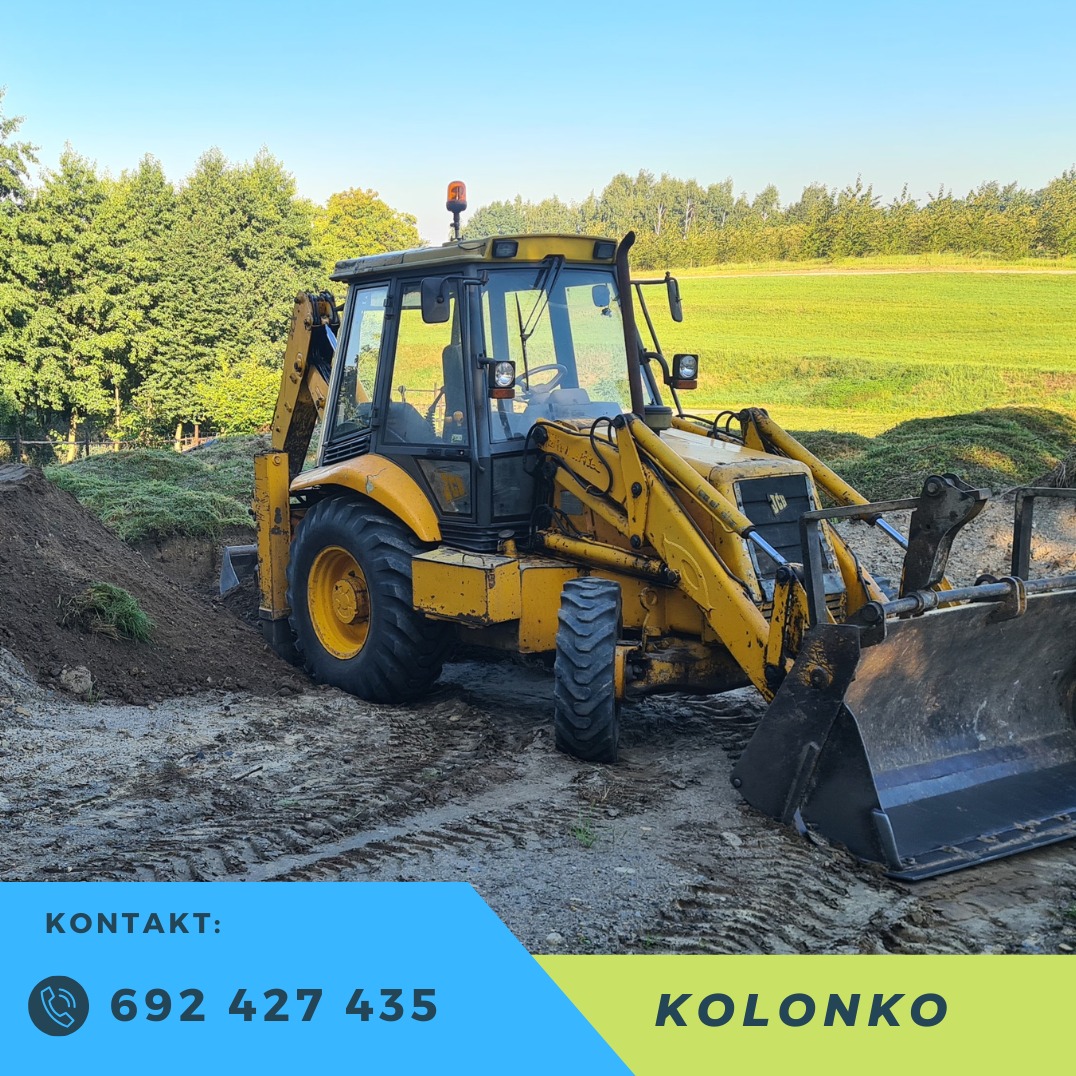 Żółta koparko-ładowarka JCB na placu budowy, świeżo wykopana ziemia, w tle zielone drzewa i łąka. Widoczne ślady opon na ubitej ziemi.