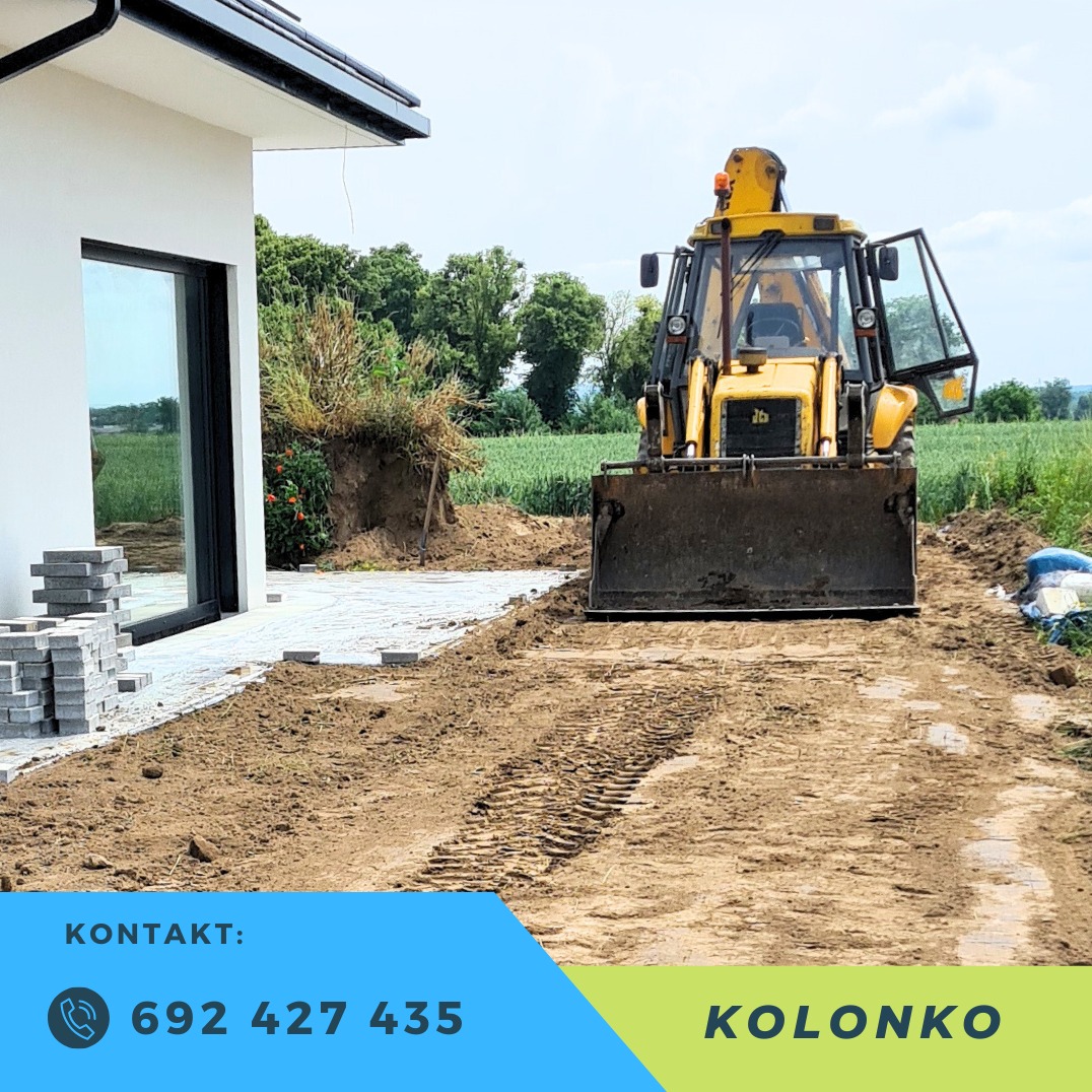 Żółta koparko-ładowarka podczas niwelacji terenu pod kostkę brukową w Raciborzu. Ślady opon na ziemi, budynek w tle. Częściowo ułożona kostka.