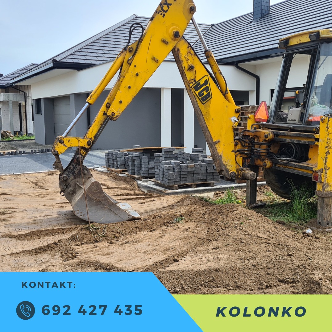 Żółta koparko-ładowarka JCB na placu budowy, obok sterty kostki brukowej. W tle dom jednorodzinny z szarą elewacją i brukowany podjazd.