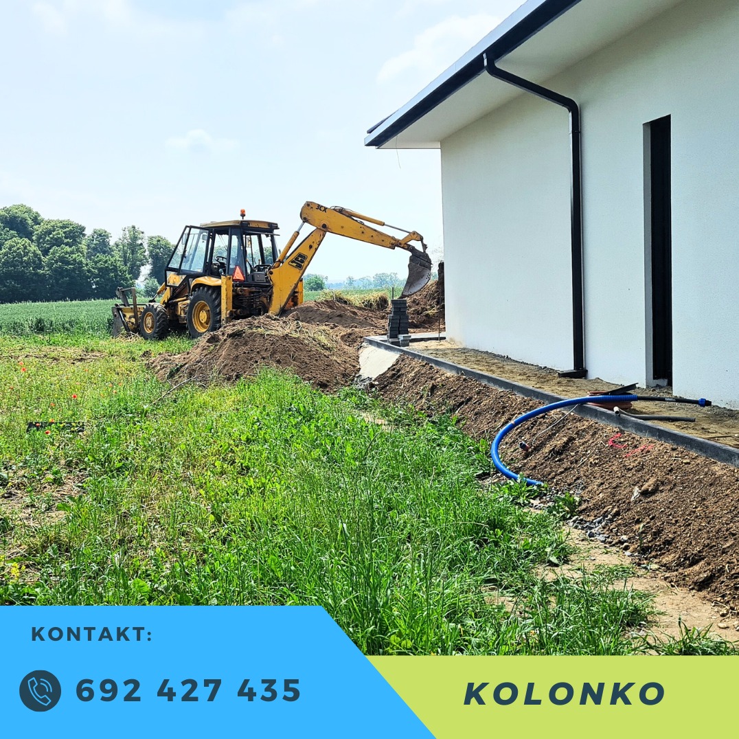 Żółta koparko-ładowarka JCB przy białym budynku z czarnymi rynnami. W tle zielone pola i las. Wykopany rów wzdłuż fundamentów, widoczny niebieski wąż.