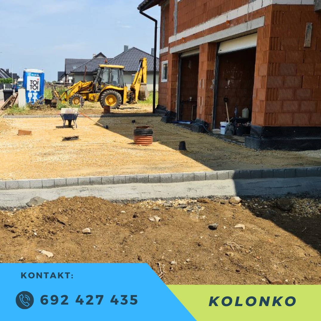 Żółta koparko-ładowarka na placu budowy, obok budynek w trakcie wznoszenia z czerwonej cegły. Widoczny fragment utwardzonej drogi z kostki.