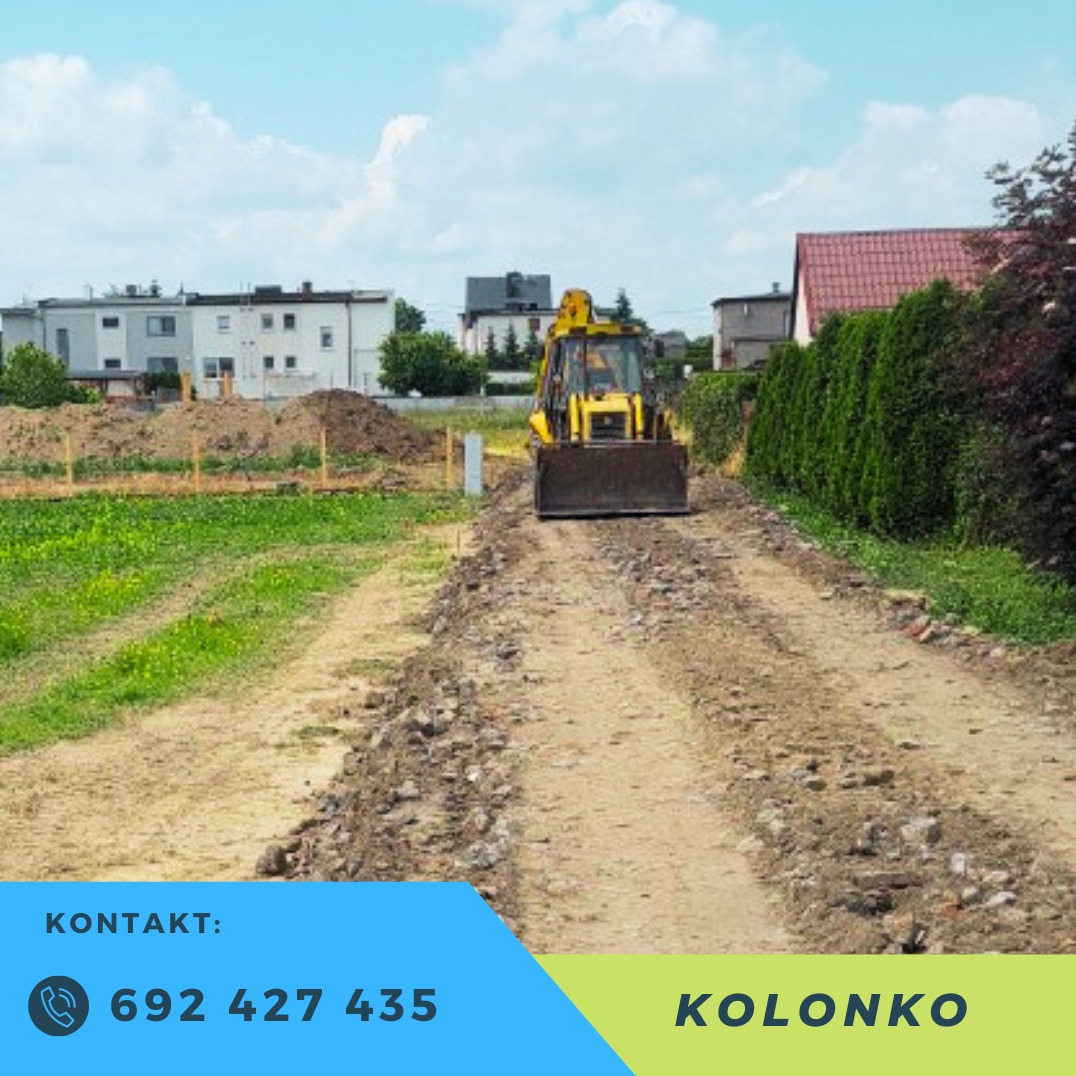 Żółta koparko-ładowarka na gruncie z gruzem i ziemią, w tle zabudowa mieszkalna i zieleń. Ślady opon na drodze wskazują na utwardzanie terenu.