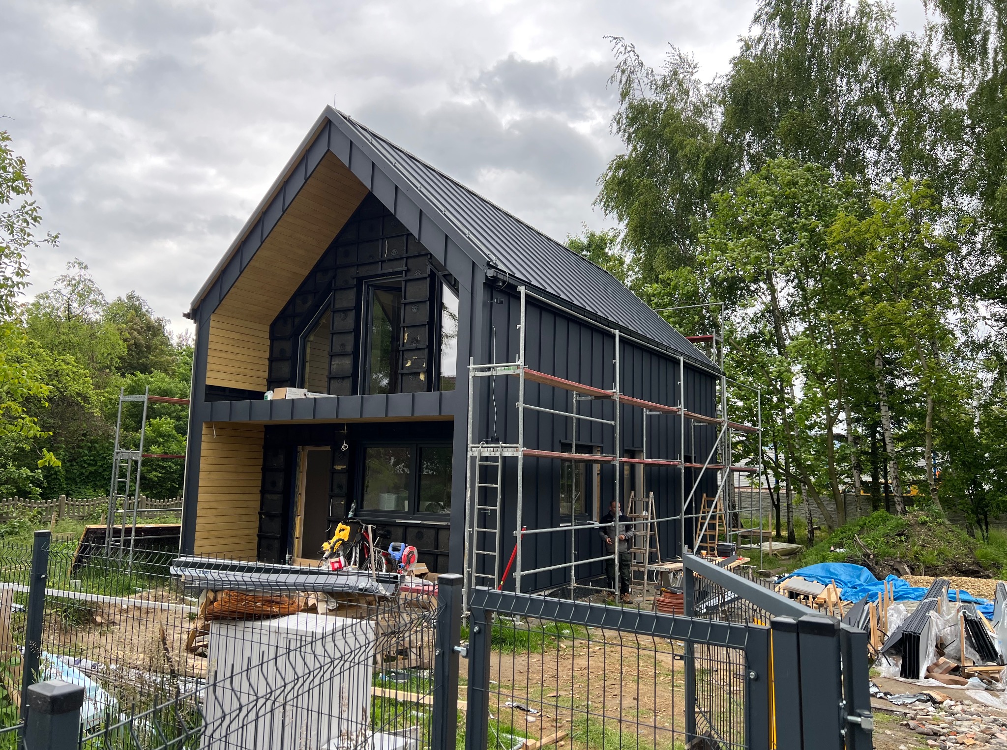 Nowoczesny dom szkieletowy w trakcie budowy, z czarną metalową elewacją i drewnianymi elementami wykończeniowymi. Widoczne rusztowania i teren budowy.