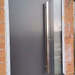 SKIBEN Skiba Przemysław - Nowoczesne, grafitowe drzwi zewnętrzne z długim, stalowym uchwytem i bocznym przeszkleniem, osadzone w surowym murze z widoczną izolacją.