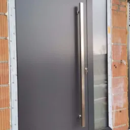 Nowoczesne, grafitowe drzwi zewnętrzne z długim, stalowym uchwytem i bocznym przeszkleniem, osadzone w surowym murze z widoczną izolacją.