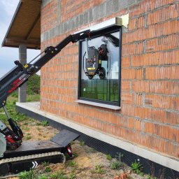 SKIBEN Skiba Przemysław - Czerwona ściana budynku w trakcie budowy z zamontowanym oknem. Do montażu użyto specjalistycznej maszyny z przyssawkami.