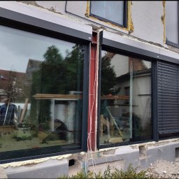 SKIBEN Skiba Przemysław - Montaż okien z roletami zewnętrznymi. Widoczne narzędzia pracy i ekipa montażowa. Okna w kolorze grafitowym, budynek w trakcie prac wykończeniowych.