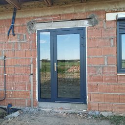 SKIBEN Skiba Przemysław - Montaż dwuskrzydłowych drzwi balkonowych w surowym murze. Widoczne kable instalacji i fragment okna. Tło to zielona łąka i hałda ziemi.