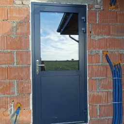 SKIBEN Skiba Przemysław - Szare drzwi z szybą zamontowane w surowym murze z czerwonej cegły. W szybie odbija się krajobraz.