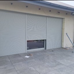 SKIBEN Skiba Przemysław - Zamknięte rolety zewnętrzne w kolorze szarym na oknie. Fragment otwartej rolety. Widoczne narzędzia montażowe i fragment budynku z szarą kostką.