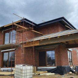 SKIBEN Skiba Przemysław - Budowa domu w trakcie: murowane ściany, okna z brązowymi ramami, rusztowania, drewniana konstrukcja dachu. Widok na budynek w surowym stanie.