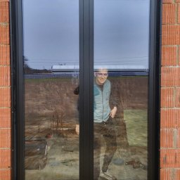 SKIBEN Skiba Przemysław - Czarne, dwuskrzydłowe okno z roletą zewnętrzną w surowym murze. W szybie odbija się uśmiechnięta osoba i narzędzia budowlane.